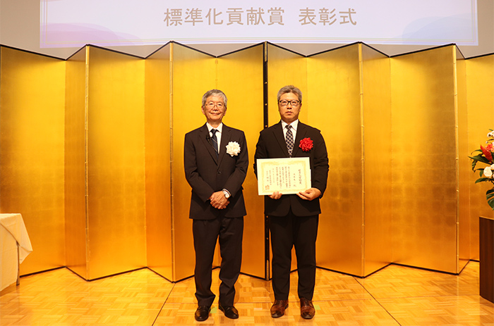 一般財団法人日本規格協会 理事長 朝日 弘 様（写真左）と受賞者の当機構 浅井 秀一（写真右）
